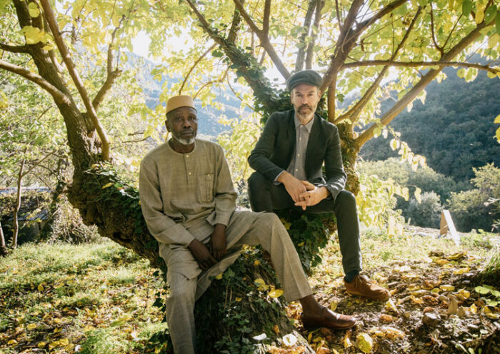 Foto de Piers Faccini y Ballaké Sissoko sentados sobre un árbol en el campo.