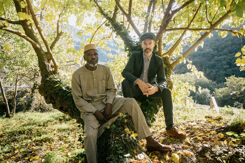 Foto de Piers Faccini y Ballaké Sissoko sentados sobre un árbol en el campo.