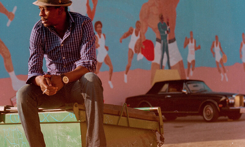 Seu Jorge – The Other Side. Portada del disco. Foto de Seu sentado sobre un contenedor de basura con un coche detrás.