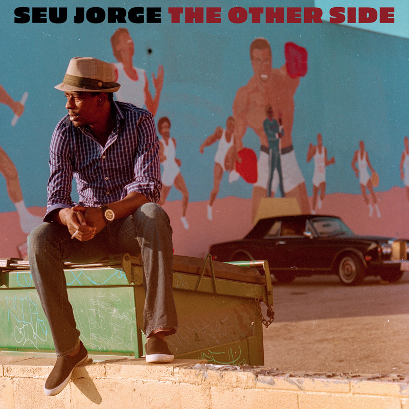 Seu Jorge – The Other Side. Portada del disco. Foto de Seu sentado sobre un contenedor de basura con un coche detrás.